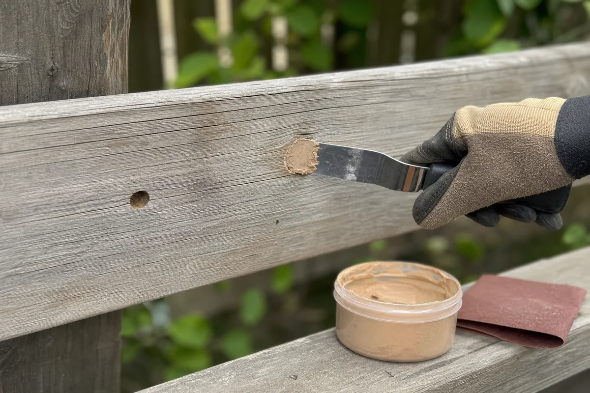 Exterior wood filler and putty knife beside an inactive carpenter bee hole in a solid fence rail