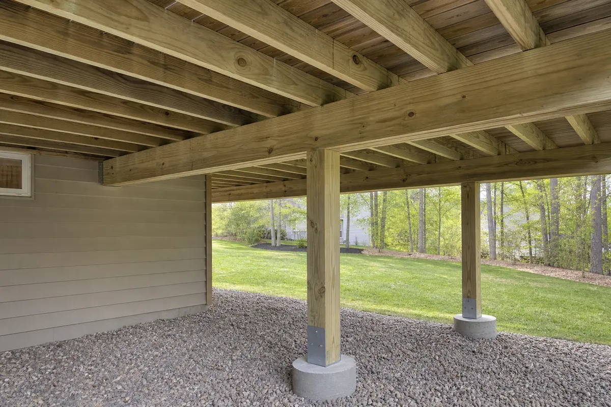 Underside of an attached wood deck showing parallel joists bearing on a continuous beam supported by posts and concrete footings