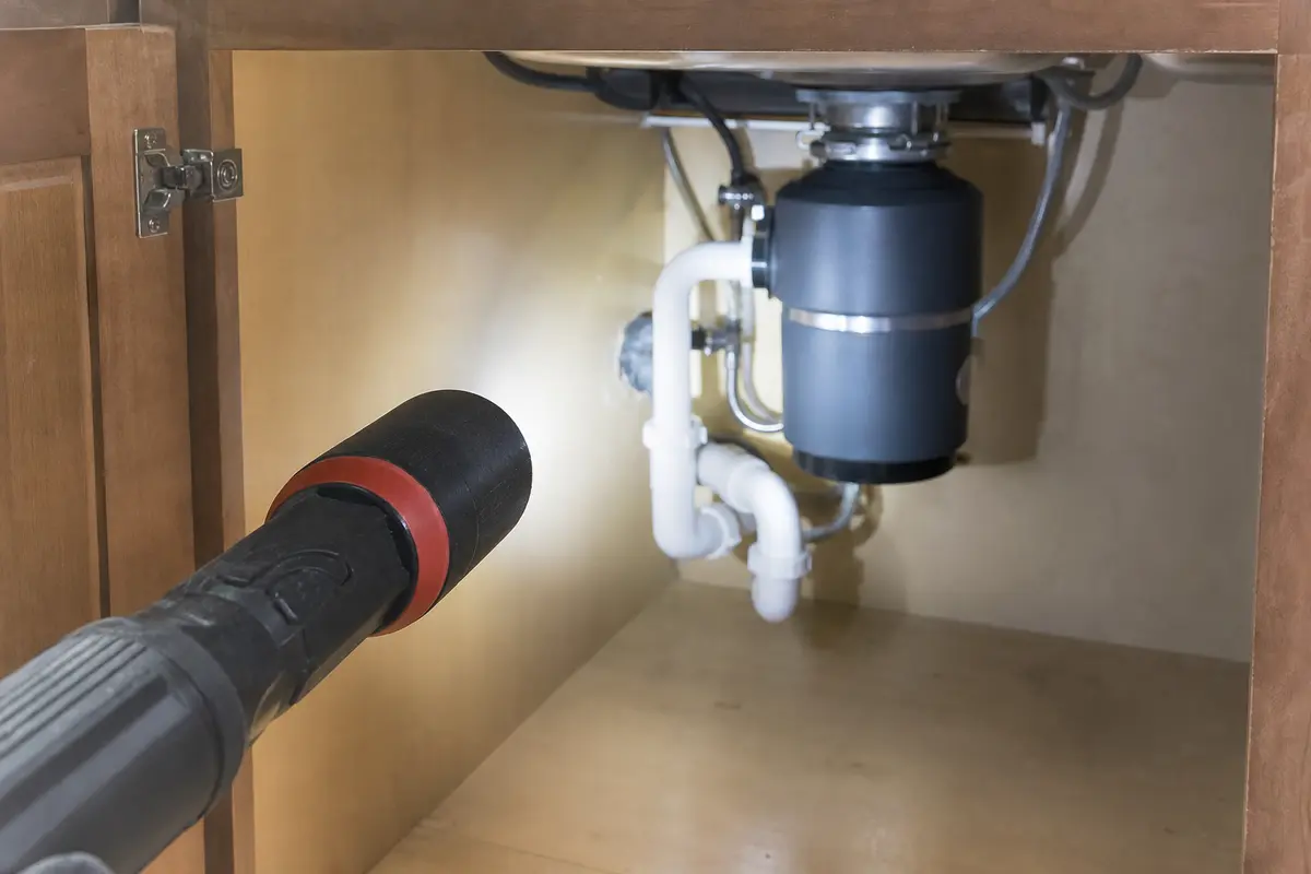 Inspection flashlight aimed into an under-sink cabinet with a garbage disposal